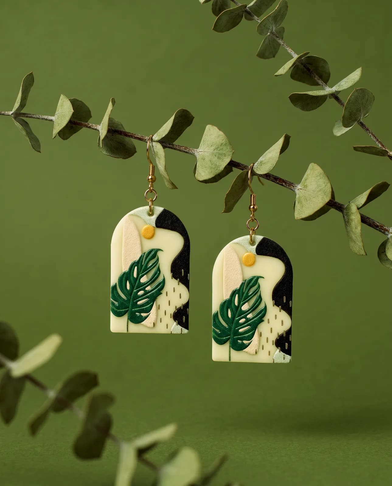 Monstera-leaf earrings styled with eucalyptus on a green backdrop